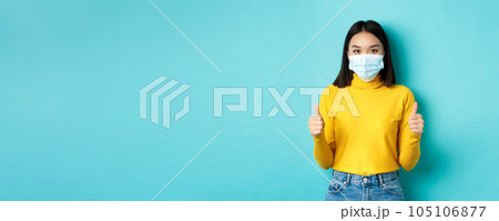 Covid-19, social distancing and pandemic concept. Supportive asian woman recommend promotion, wearing medical mask and showing thumbs up, standing over blue background 105106877
