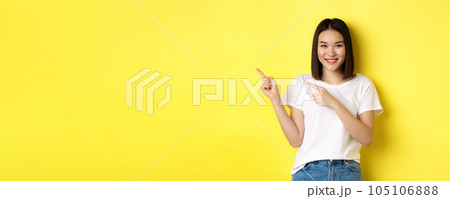 Beauty and fashion concept. Beautiful asian woman in white t-shirt pointing fingers left, standing over yellow background Beauty and fashion concept. Beautiful asian woman in white t-shirt pointing fingers left, standing over yellow background 105106888