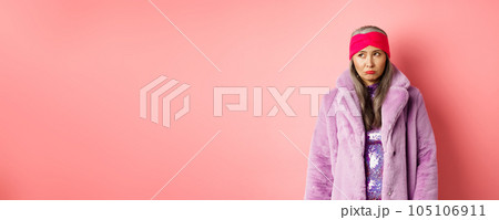 Sad and indecisive asian senior woman in fashionable faux fur coat looking left and sulking upset, standing against pink background 105106911
