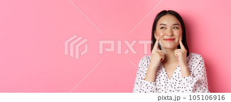 Beauty and skin care concept. Close-up of beautiful asian woman poking cheeks and smiling satisfied, standing over pink background Beauty and skin care concept. Close-up of beautiful asian woman poking cheeks and smiling satisfied, standing over pink background 105106916