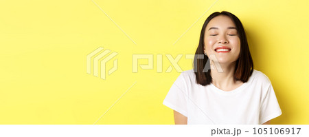 Close up of happy and relaxed asian woman enjoying sun, smiling with eyes closed and looking joyful, standing over yellow background Close up of happy and relaxed asian woman enjoying sun, smiling with eyes closed and looking joyful, standing over yellow background 105106917