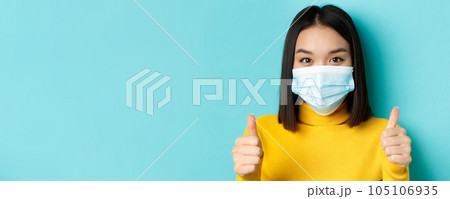 Covid-19, social distancing and pandemic concept. Close up of young asian woman in medical mask showing thumbs up, say yes, praise good offer, standing over blue background 105106935