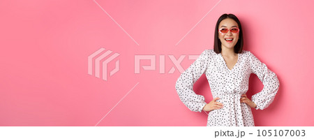 Tourism and lifestyle concept. Beautiful korean woman on vacation, wearing sunglasses and dress, looking left with happy smile, standing over pink background Tourism and lifestyle concept. Beautiful korean woman on vacation, wearing sunglasses and dress, looking left with happy smile, standing over pink background 105107003