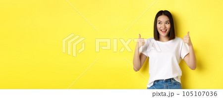 Pretty young asian woman in white t-shirt, showing thumbs up and smiling, praise good offer, recommend product, standing satisfied over yellow background Pretty young asian woman in white t-shirt, showing thumbs up and smiling, praise good offer, recommend product, standing satisfied over yellow background 105107036