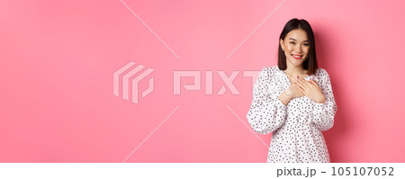 Thankful Korean girl in dress smiling, holding hands on heart and looking grateful at camera, touched with nice gesture, standing over pink background Thankful Korean girl in dress smiling, holding hands on heart and looking grateful at camera, touched with nice gesture, standing over pink background 105107052