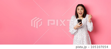 Online shopping and beauty concept. Excited asian woman winning in internet, holding smartphone and rejoicing, smiling happy and celebrating, standing over pink background 105107054