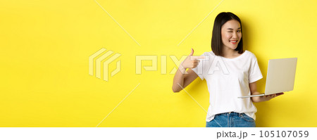 E-commerce and shopping concept. Good-looking asian girl winking at camera, smiling and pointing finger a laptop, showing something, standing over yellow background 105107059