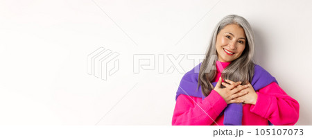 Close up of stylish asian mother holding hands on heart, looking touched and heartfelth, saying thank you, standing over white background Close up of stylish asian mother holding hands on heart, looking touched and heartfelth, saying thank you, standing over white background 105107073