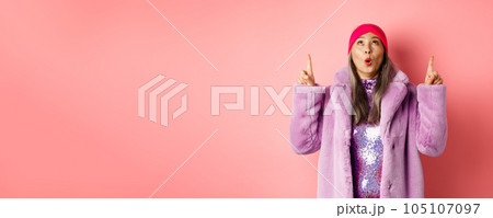 Impressed asian elderly woman in stylish fake-fur coat checking out promo, looking and pointing fingers up, standing over pink background 105107097