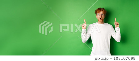Shocked and worried redhead man in glasses gasping and staring at camera, pointing fingers up at advertisement, standing over green background 105107099