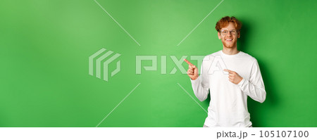 Handsome young man with red hair and beard smiling, pointing fingers left at logo, showing advertisement, standing over green background Handsome young man with red hair and beard smiling, pointing fingers left at logo, showing advertisement, standing over green background 105107100