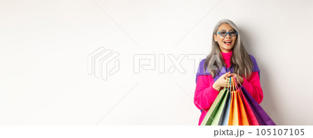 Stylish senior asian woman in sunglasses going shopping on holiday sale, holding paper bags and smiling, standing over white background Stylish senior asian woman in sunglasses going shopping on holiday sale, holding paper bags and smiling, standing over white background 105107105