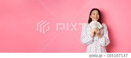 Shopping concept. Happy and satisfied asian woman winning prize money, showing dollars and rejoicing, standing over pink background 105107137