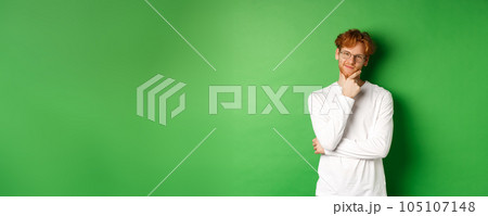 Intrigued young man with red hair, wearing glasses, looking at camera pleased and thoughtful, having an idea, standing over green background 105107148