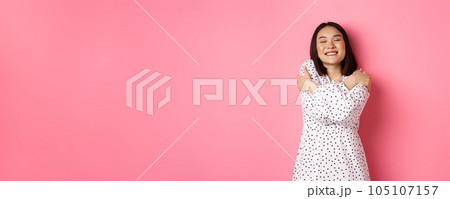 Beautiful asian woman feeling romantic, hugging herself, close eyes and smiling with dreamy face, standing happy over pink background 105107157
