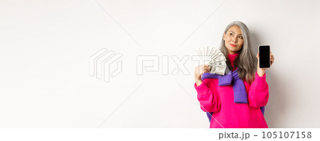 Fashionable asian senior woman showing money dollars and blank smartphone screen, demonstrate online shop, standing over white background Fashionable asian senior woman showing money dollars and blank smartphone screen, demonstrate online shop, standing over white background 105107158