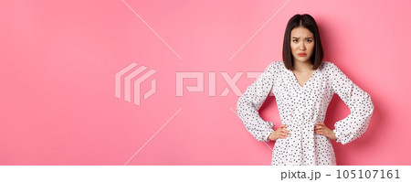 Cute offended girl frowning, holding hands on waist and staring at camera disappointed, standing upset and gloomy over pink background Cute offended girl frowning, holding hands on waist and staring at camera disappointed, standing upset and gloomy over pink background 105107161