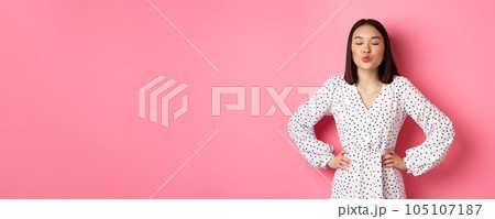 Beautiful asian woman in dress pucker lips and close eyes, waiting for kiss, standing over pink background 105107187