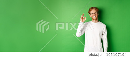 Happy male student with red hair, wearing glasses, showing okay sign in approval and smiling satisfied, standing over green background Happy male student with red hair, wearing glasses, showing okay sign in approval and smiling satisfied, standing over green background 105107194