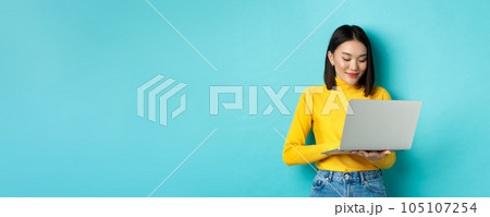Beautiful asian female student working on laptop, typing on keyboard and looking at screen with pleased smile, standing over blue background Beautiful asian female student working on laptop, typing on keyboard and looking at screen with pleased smile, standing over blue background 105107254