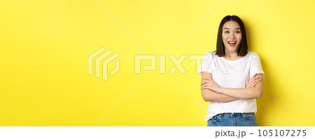 Beauty and fashion concept. Happy asian woman in white t-shirt, cross arms on chest, looking amazed at camera, standing over yellow background 105107275