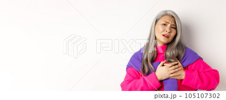 Worried asian mother holding hands on heart and grimacing, looking anxious and having pain in chest, feeling hurt, standing over white background 105107302