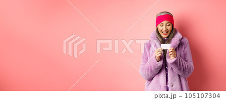 Shopping and fashion concept. Beautiful and stylish asian woman looking at plastic credit card, smiling pleased, standing over pink background Shopping and fashion concept. Beautiful and stylish asian woman looking at plastic credit card, smiling pleased, standing over pink background 105107304