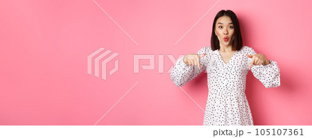 Impressed asian woman in dress pointing fingers down, look there gesture, check out discounts and sales, standing amazed over pink background 105107361