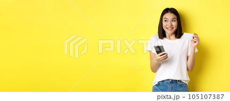 E-commerce and online shopping concept. Cheerful asian girl paying in internet, holding smartphone and plastic credit card, smiling and looking left, yellow background E-commerce and online shopping concept. Cheerful asian girl paying in internet, holding smartphone and plastic credit card, smiling and looking left, yellow background 105107387