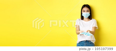 Coronavirus, quarantine and medicine concept. Cute asian girl giving medical masks and looking at camera, yellow background Coronavirus, quarantine and medicine concept. Cute asian girl giving medical masks and looking at camera, yellow background 105107399