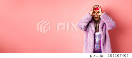 Fashion and shopping. Amazed asian senior woman checking out awesome valentines day promotion, looking through hands binoculars and gasping impressed, pink background 105107405