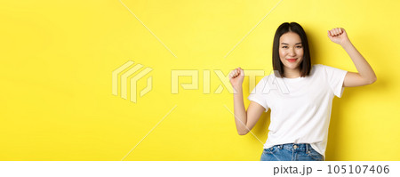 Carefree asian girl dancing and having fun, posing in white t-shirt against yellow background 105107406