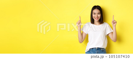 Beauty and fashion concept. Beautiful asian woman in white t-shirt pointing fingers up, standing over yellow background Beauty and fashion concept. Beautiful asian woman in white t-shirt pointing fingers up, standing over yellow background 105107410