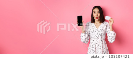 Cute asian woman shopping online, showing bank credit card and mobile screen, smiling and looking left at copy space, standing over pink background 105107421