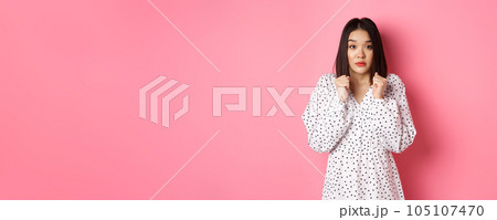 Scared cute asian girl in dress looking worried, feeling frightened and staring at camera nervously, standing over pink background Scared cute asian girl in dress looking worried, feeling frightened and staring at camera nervously, standing over pink background 105107470