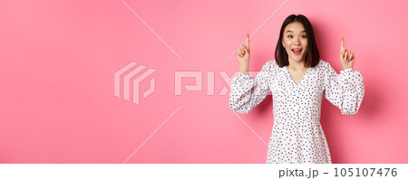 Amazed brunette asian girl pointing fingers up, showing advertisement, demonstrate spring promo offer, standing over pink background 105107476