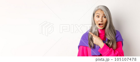 Close-up of amazed and intrigued asian senior woman pointing finger left at something cool, staring at camera impressed, standing over white background 105107478