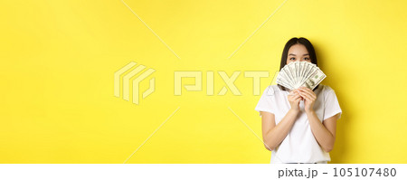 Cute asian woman hiding face behind money, peeking at camera satisfied, earn cash, standing over yellow background Cute asian woman hiding face behind money, peeking at camera satisfied, earn cash, standing over yellow background 105107480