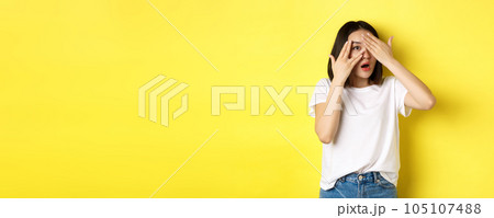 Intrigued asian girl waiting for surprise, peeking through fingers on eyes, smiling happy, standing over yellow background Intrigued asian girl waiting for surprise, peeking through fingers on eyes, smiling happy, standing over yellow background 105107488