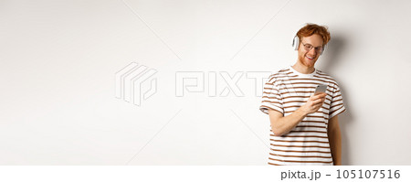 Technology concept. Young redhead hipster in glasses, listening music in headphoes, reading message on smartphone, white background Technology concept. Young redhead hipster in glasses, listening music in headphoes, reading message on smartphone, white background 105107516