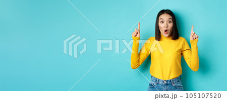 Shopping concept. Impressed young korean woman showing advertisement on top, pointing fingers up and gasping amazed, saying wow, standing over blue background 105107520