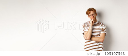 Young man with red messy hair and glasses looking doubtful, thinking and squinting at camera, standing thoughtful over white background Young man with red messy hair and glasses looking doubtful, thinking and squinting at camera, standing thoughtful over white background 105107548