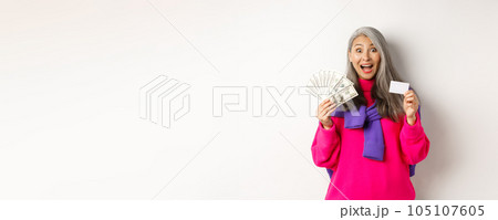 Shopping concept. Amazed asian granny showing plastic credit card and money dollars, want to buy something, standing over white background Shopping concept. Amazed asian granny showing plastic credit card and money dollars, want to buy something, standing over white background 105107605
