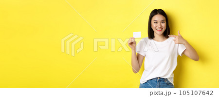 Young asian woman in casual white t-shirt showing plastic credit card and smiling at camera, yellow background 105107642