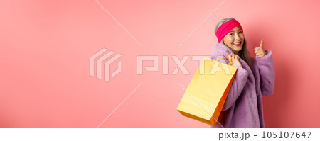 Stylish asian senior woman going shopping, carry paper bag on shoulder and showing thumb-up, recommending store discounts, pink background 105107647