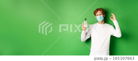 Covid-19, virus and social distancing concept. Smiling young man with red hair, wearing face mask from coronavirus, showing okay sign and hand sanitizer, green background 105107663