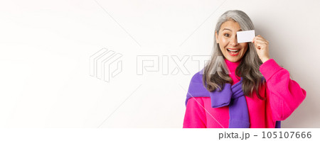 Shopping concept. Stylish asian senior woman smiling and showing plastic credit card, paying contactless, standing over white background Shopping concept. Stylish asian senior woman smiling and showing plastic credit card, paying contactless, standing over white background 105107666