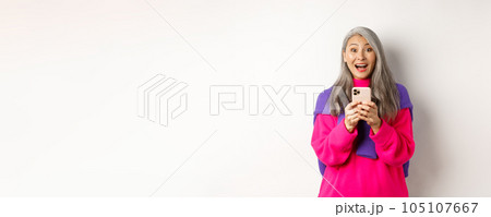 Surprised asian woman smiling at camera after reading promotion on smartphone, standing with mobile phone over white background Surprised asian woman smiling at camera after reading promotion on smartphone, standing with mobile phone over white background 105107667