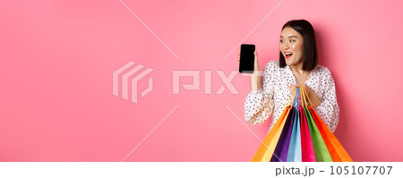 Attractive asian woman showing smartphone app and shopping bags, buying online via application, standing over pink background Attractive asian woman showing smartphone app and shopping bags, buying online via application, standing over pink background 105107707