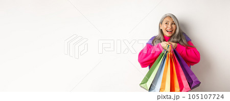 Fashionable senior asian woman shopaholic, hugging shopping bags and smiling joyful, buying with discounts, standing over white background 105107724